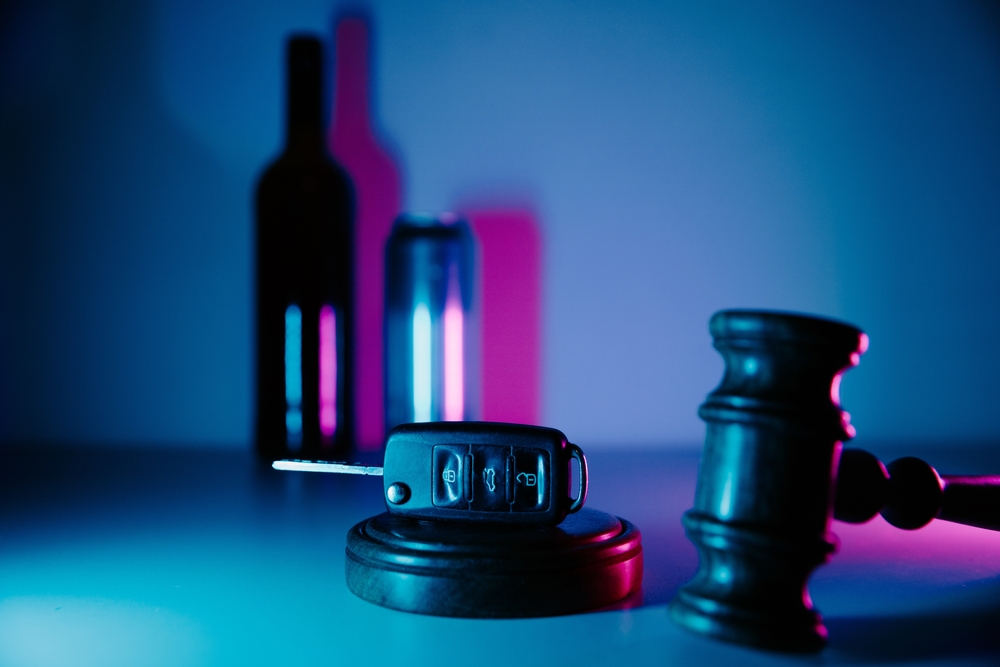 A car key fob on a pedestal next to a judge’s gavel, with blurred alcohol bottles and cans in the background under blue and pink lighting.